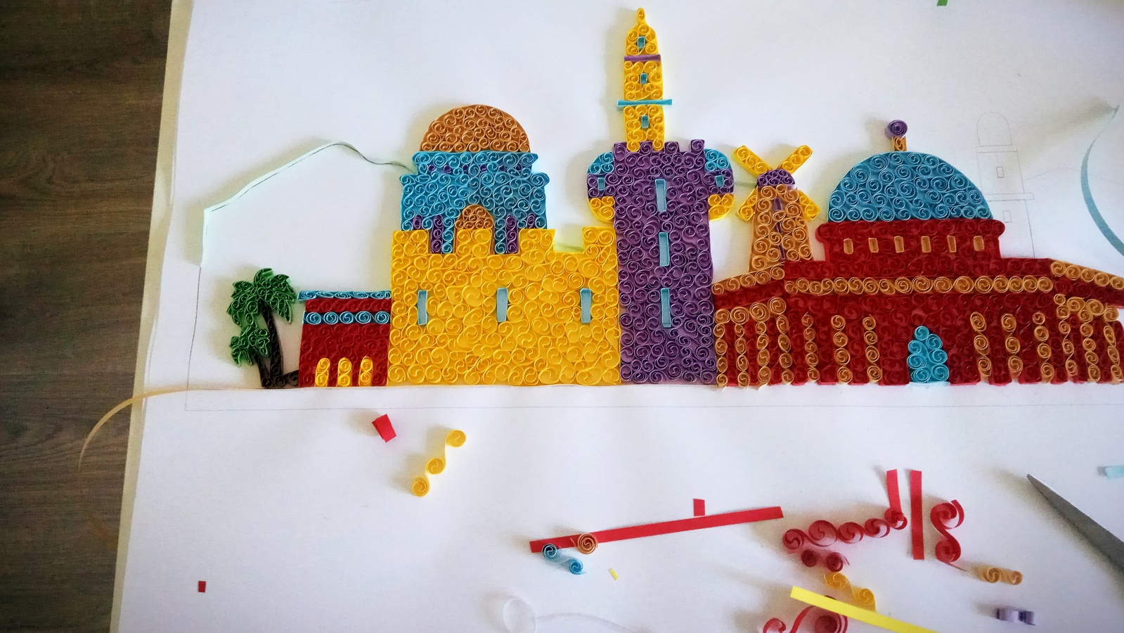 A colorful paper quilling artwork depicts a cityscape with domed and towered buildings on a white background, surrounded by strips of paper and quilling tools.