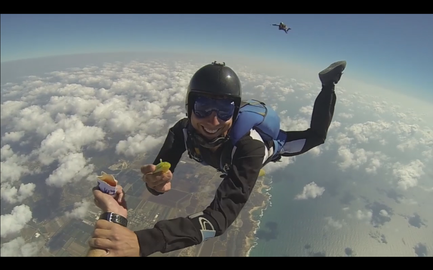 A man skydiving in the middle of the air, above clouds, holds an apple dipped in honey as he smiles facing the camer.a