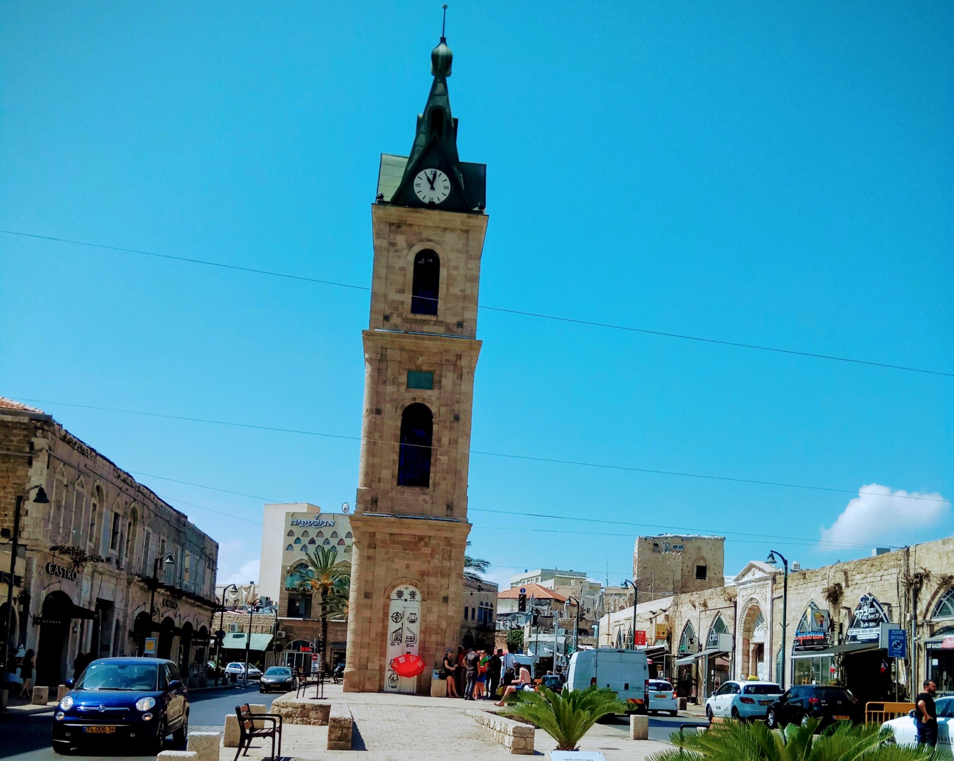 Jaffa Clock Tower photo by Eli Halfin The tall Jaffa clock tower stands in a bustling square under a clear blue sky. People gather near the base, surrounded by historic buildings. Cars are parked along the street, and palm plants line the foreground.
