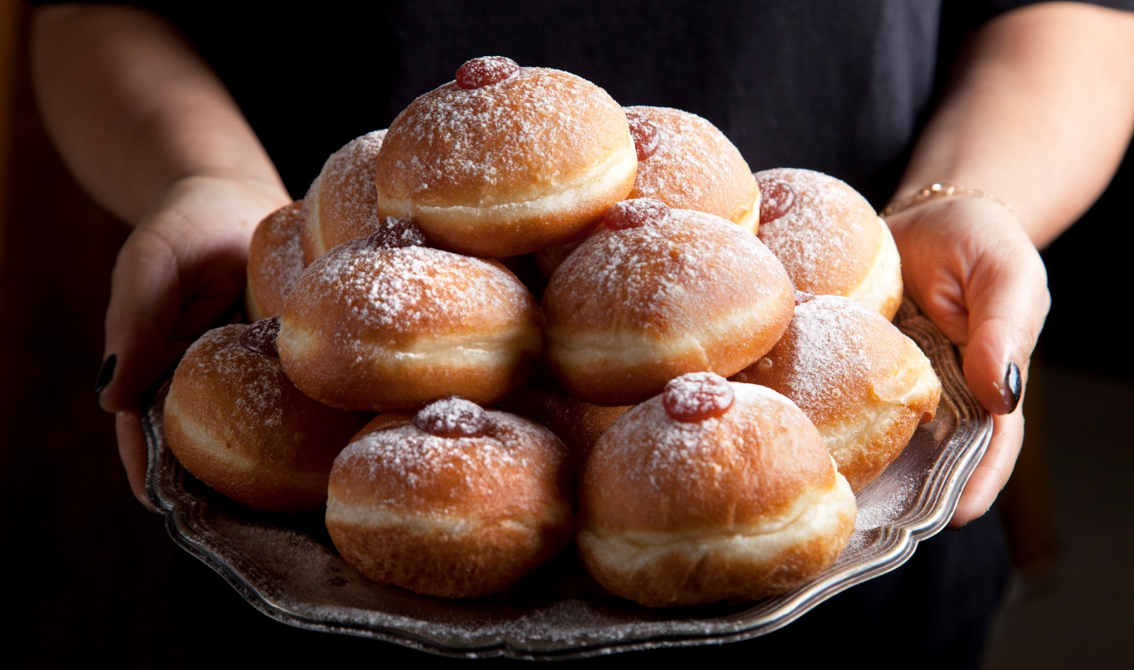 Lehamim Bakery sufganiyot in classic flavors. Photo by Daniel Layla A person holds a large silver tray filled with jelly doughnuts topped with powdered sugar, each with a bit of red jelly visible in the center.