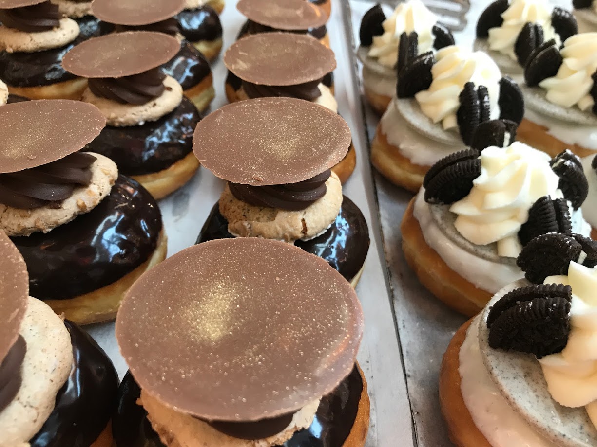 Rows of gourmet donuts topped with chocolate glaze, cookies, swirls of cream, chocolate discs, and mini Oreo cookie pieces.