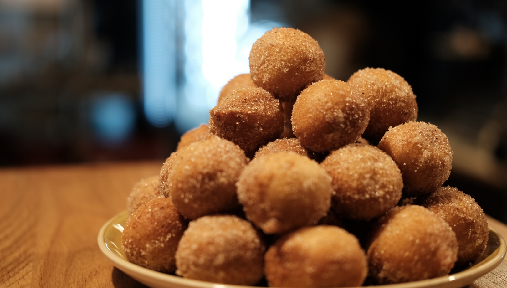 Lechem Borkin Cake Donut Holes. Photo by Eran Borkin A plate stacked high with round, sugar-coated doughnut holes sits on a wooden surface, with a blurred background.