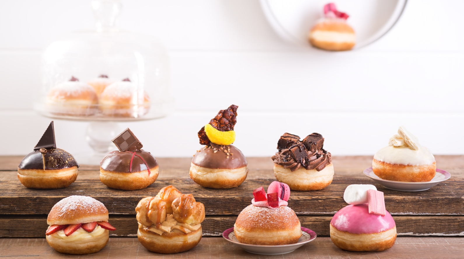 Metuka Bakery sufganiyot. Photo by Benny Gam Zu Letova An assortment of decorative gourmet donuts with various toppings, including chocolate pieces, fruit, icing, and candies, arranged on wooden boards with more donuts in the background and on a wall-mounted plate.