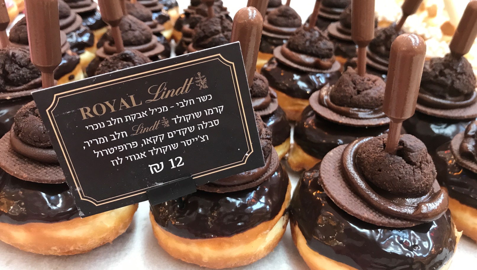 Roladin’s Royal Lindt sufganiyot. Photo by Nicky Blackburn Rows of gourmet chocolate donuts topped with glossy chocolate glaze, a chocolate cookie, and a plastic pipette filled with chocolate sauce. A sign in Hebrew and English displays the Lindt brand and the price of 12 shekels.