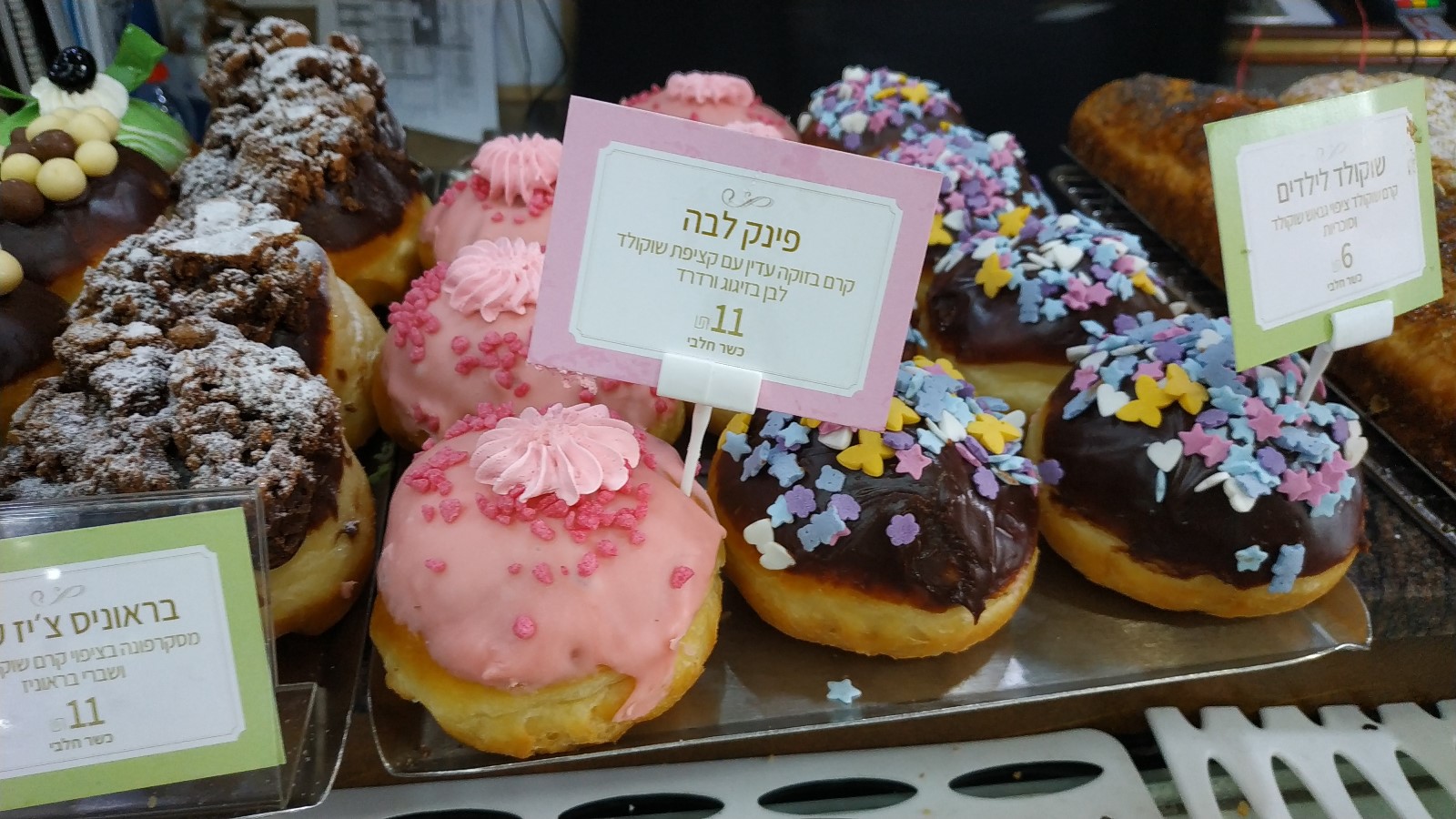 Shany Bakery’s sufganiyot. Photo by Jessica Halfin Assorted festive donuts on trays, some topped with pink or chocolate icing and colorful sprinkles, displayed with Hebrew signs labeling flavors and prices.
