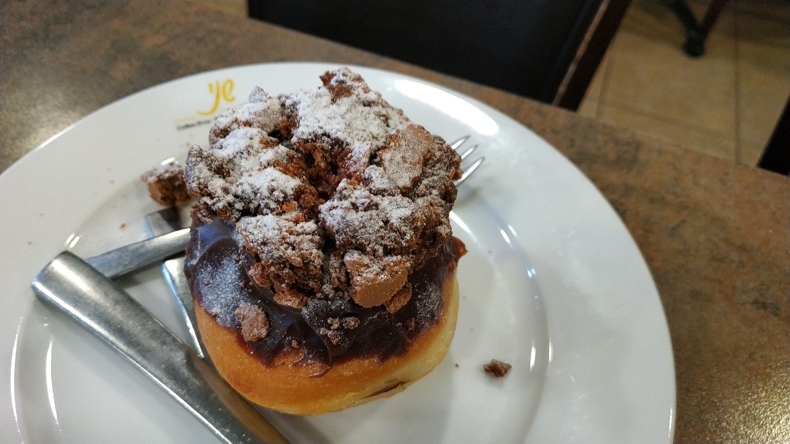 Brownie-Cheesecake donut at Shany Bakery. Photo by Jessica Halfin A chocolate-covered donut topped with crumbled chocolate and powdered sugar sits on a white plate with a fork and knife on a brown table.