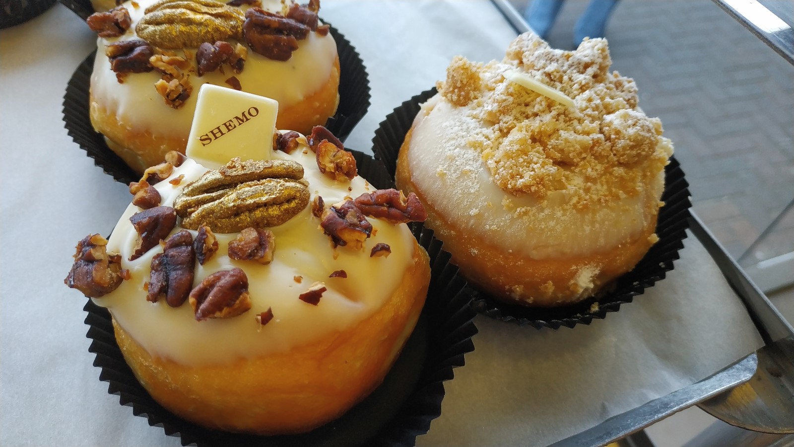 Shemo Bakery sufganiyot. Photo by Jessica Halfin Three gourmet donuts sit on a tray; two are topped with pecans and a white chocolate square labeled SHEMO, while the third has a crumbly streusel topping and a white chocolate piece.