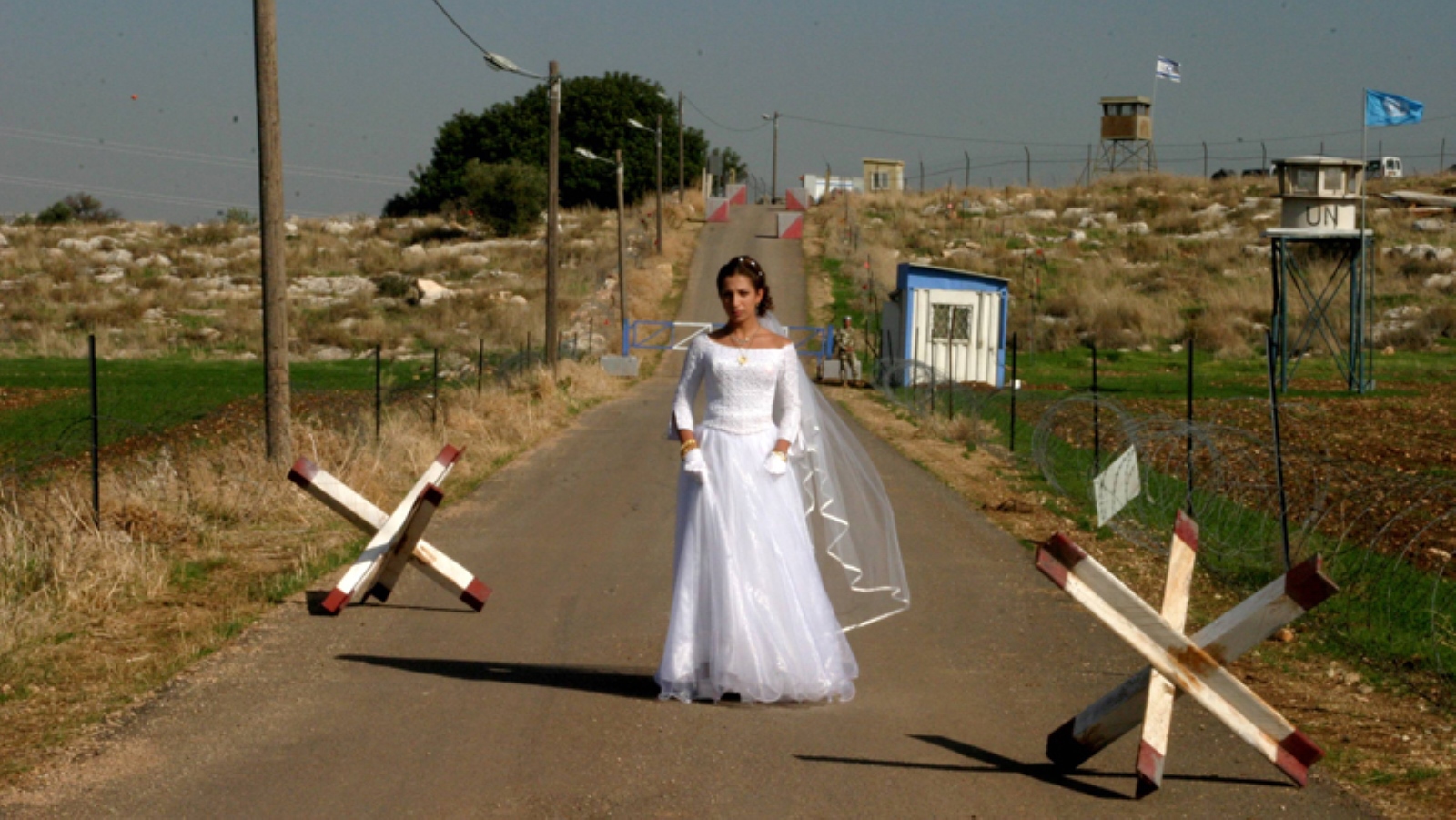 Video still from the film The Syrian Bride featuring a bride in a white wedding dress standing confidently on a deserted road flanked by wooden barriers. The background features a rural landscape with dry grass, a small building, and a watchtower under a clear blue sky. 