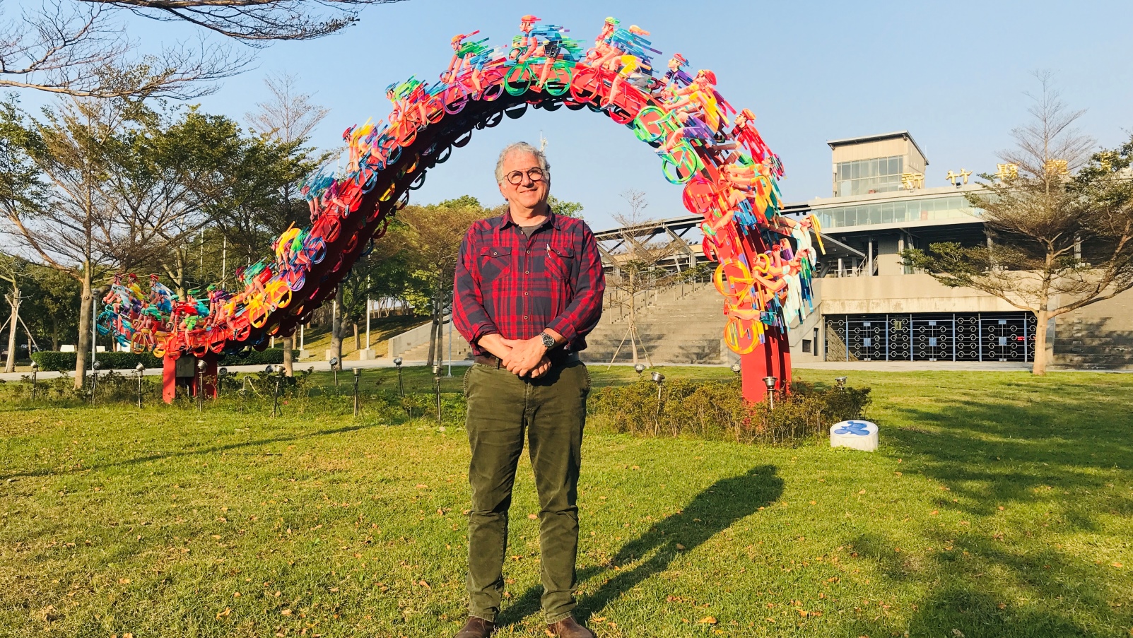 David Gerstein stands in green pants and a red plaid shirt, smiling on green grass in front of a colorful sculpture made of bicycles arranged in an arch. Trees and a modern building are in the background under a clear blue sky.