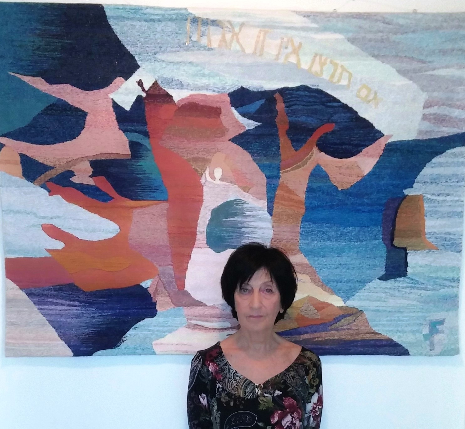 Silvia Feinstein, with short dark hair, stands in front of a colorful abstract tapestry with a mix of blue, pink, and orange hues. The tapestry features abstract forms and text at the top. She wears a black floral-patterned blouse.
