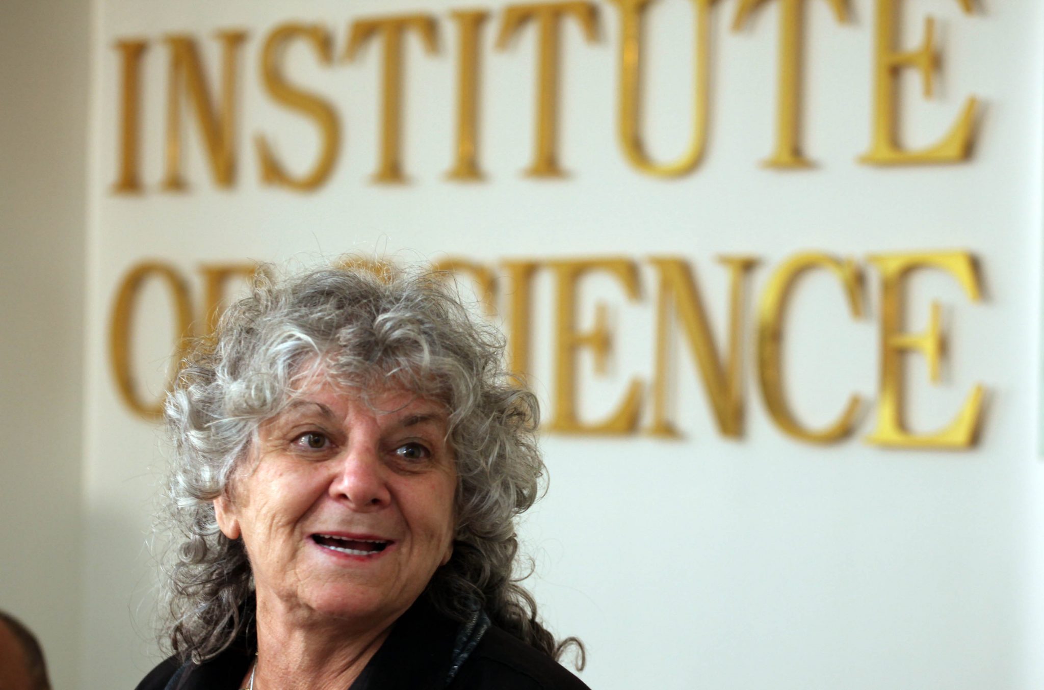 Ada Yonath, with grey curly hair, smiles while standing in front of a wall with large gold letters reading INSTITUTE OF SCIENCE. 
