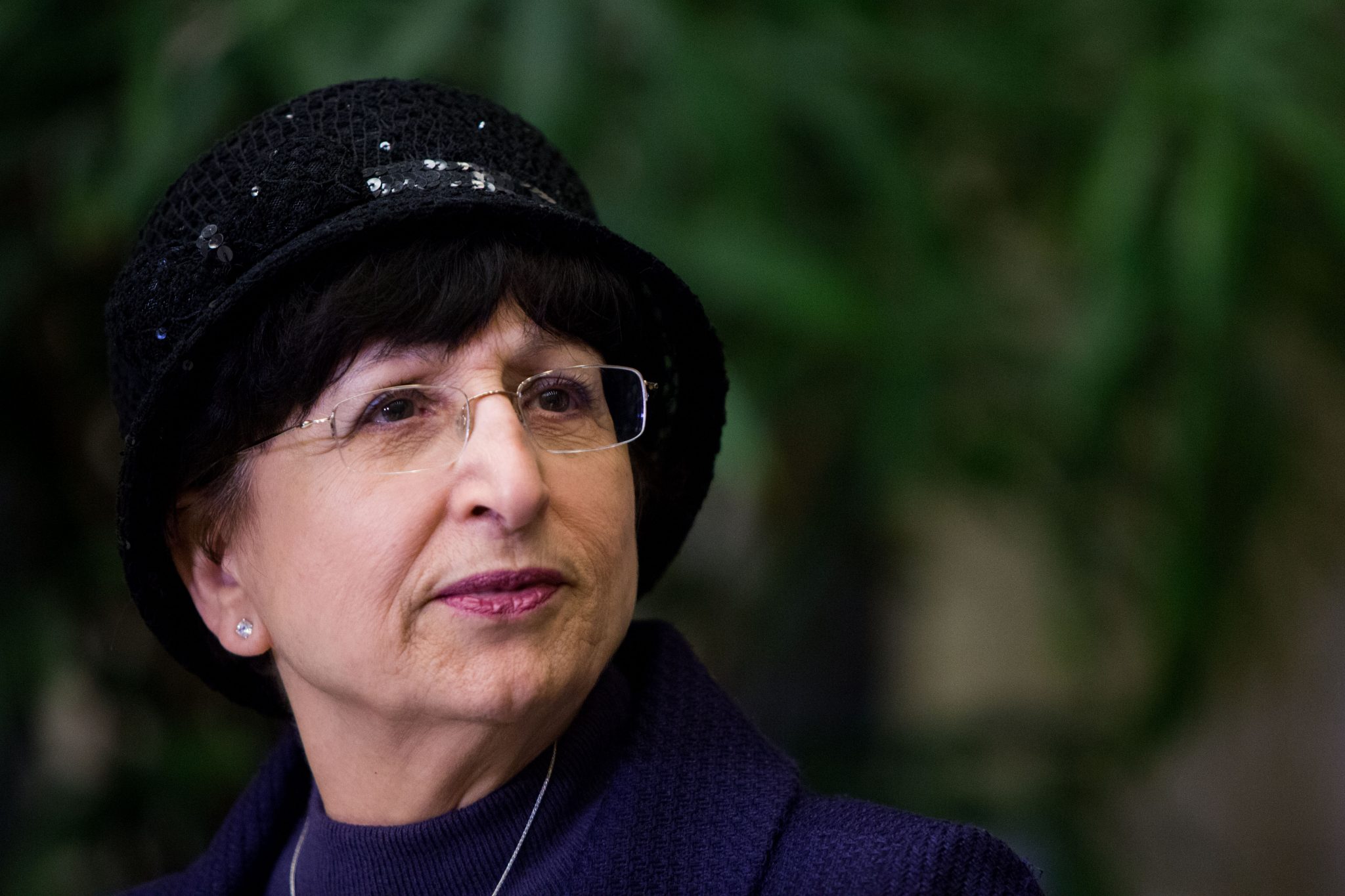 Adina Bar-Shalom wears glasses and a dark hat with a purple coat, looking thoughtfully to the side. The background is blurred with green foliage. 