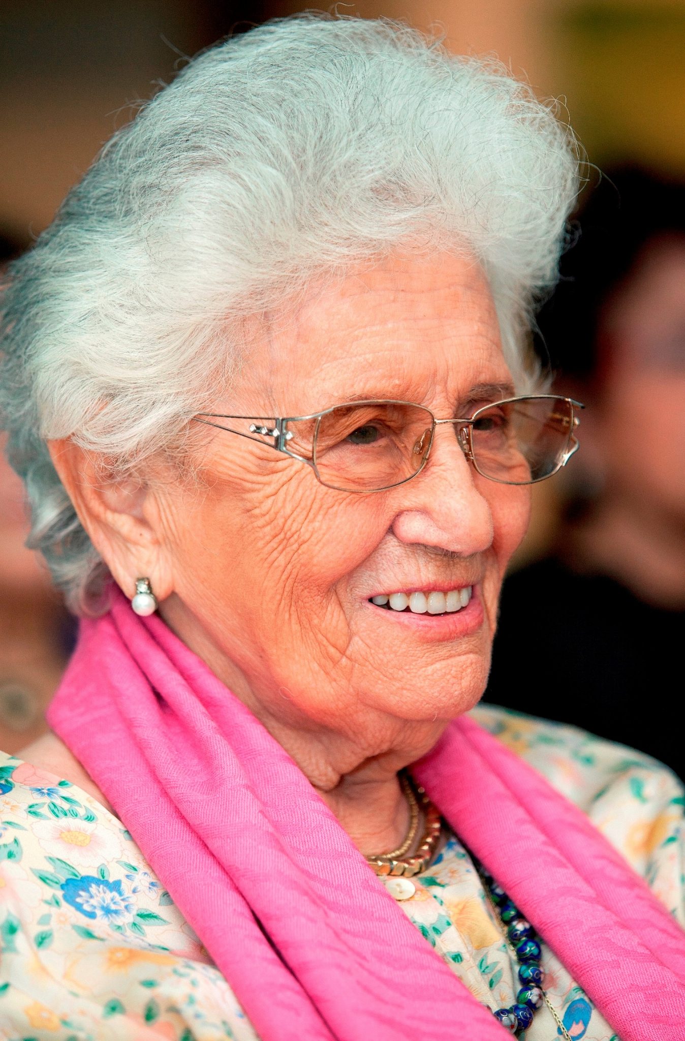 Ruth Dayan with white hair and glasses wearing a floral blouse and a pink scarf. She is smiling and looking to the side.