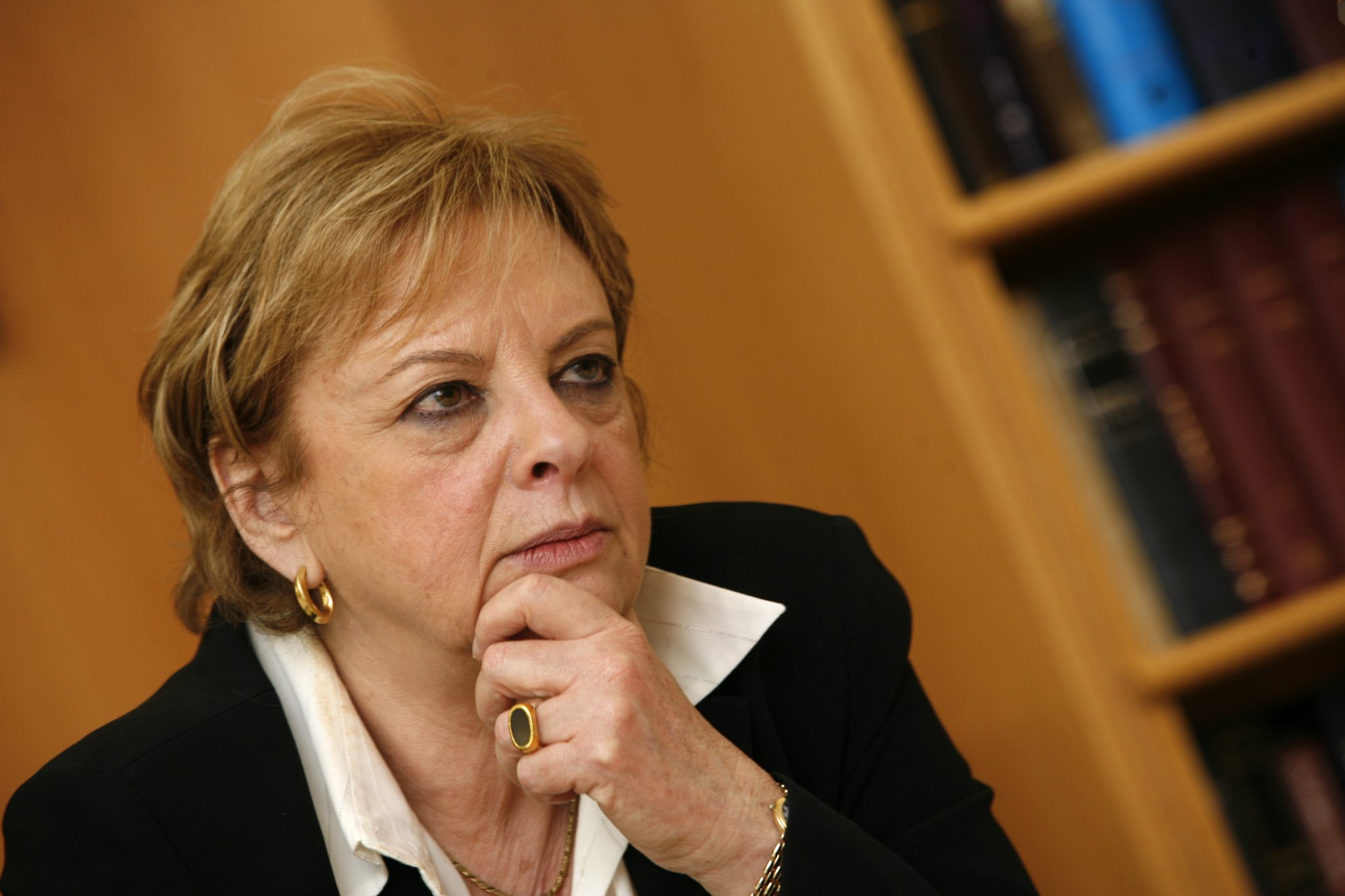 Dorit Beinisch sits with short hair and gold hoop earrings, resting her chin on her hand. She is wearing a black blazer and a white shirt. Bookshelves are visible in the background. 