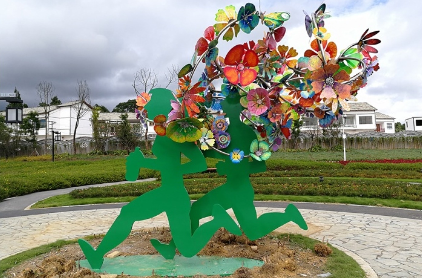 A vibrant outdoor sculpture features two green running figures with colorful, abstract flowers flowing behind them. The background includes a path, greenery, and cloudy skies.
