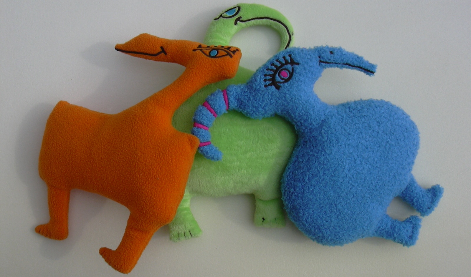 Three plush toy creatures lie on a surface. The toys are uniquely shaped and colored: one is orange, another green, and the last is blue. They have exaggerated features and playful designs, each featuring stitched eyes and playful expressions.