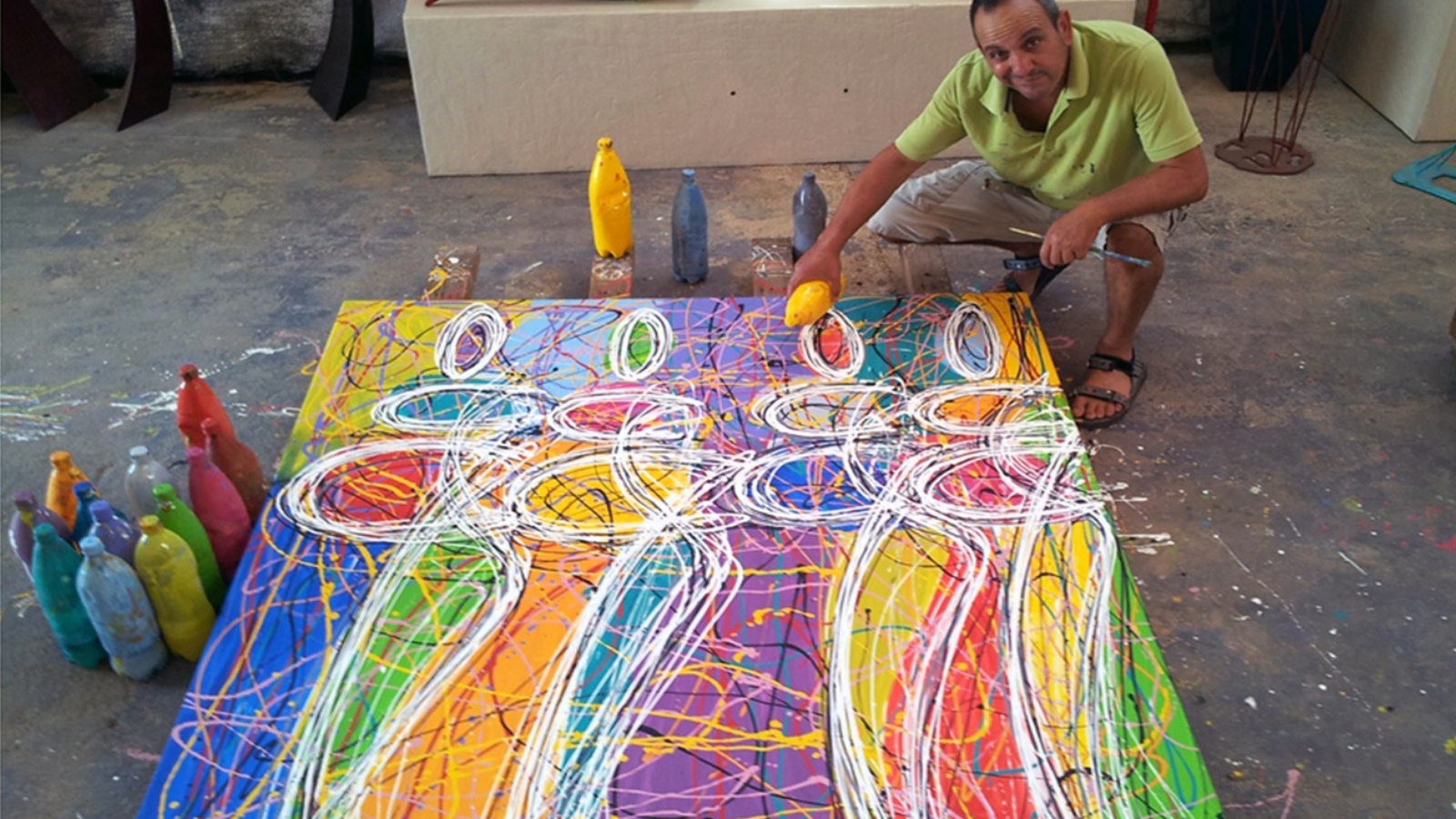 Artist Jojo Ohayon squats beside a colorful abstract painting on the floor, with vibrant figures and swirling lines. He holds a bottle and is surrounded by similar bottles in various colors. He wears a light green shirt and dark shorts.