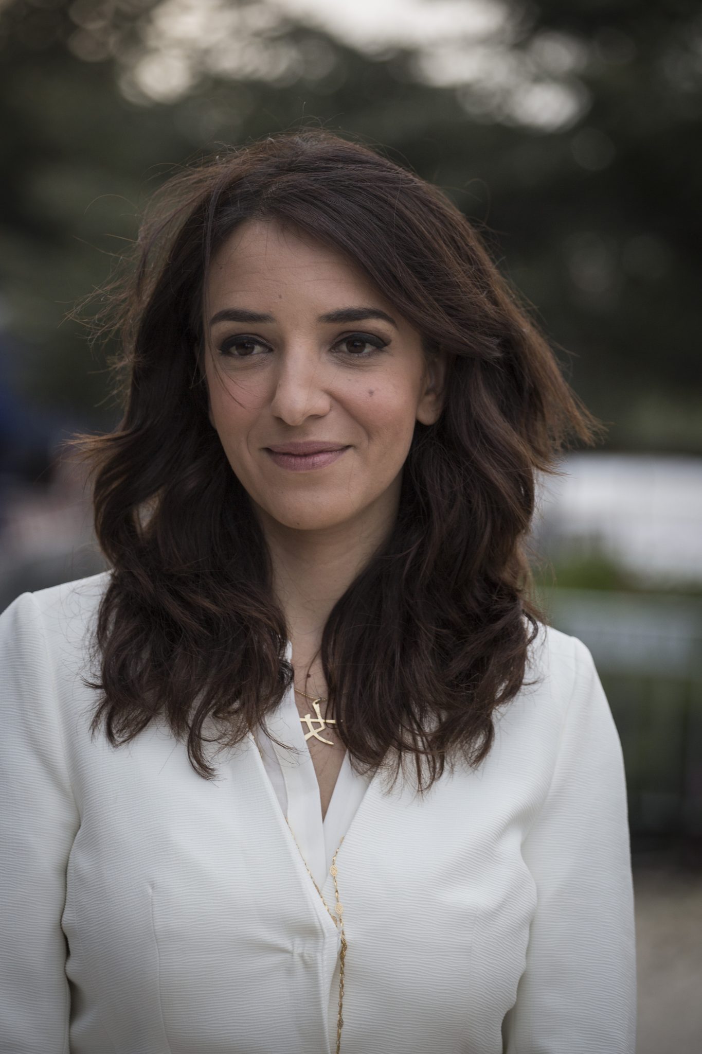 Lucy Aharish with long, wavy dark hair and wearing a white top, stands outdoors. She is smiling softly. A gold necklace with a pendant is visible and the background is blurred greenery. 