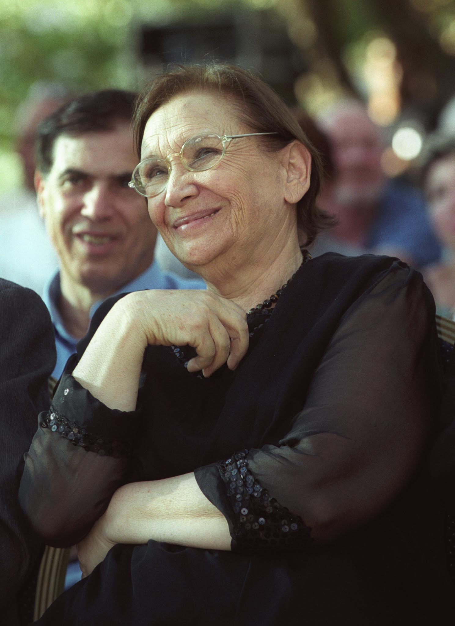 Naomi Shemer smiles with glasses on and stands amongst a crowd outdoors. She's wearing a black outfit with sheer sleeves. A man is seated behind her, with more people blurred in the background and the setting appears to be a public event or gathering. 