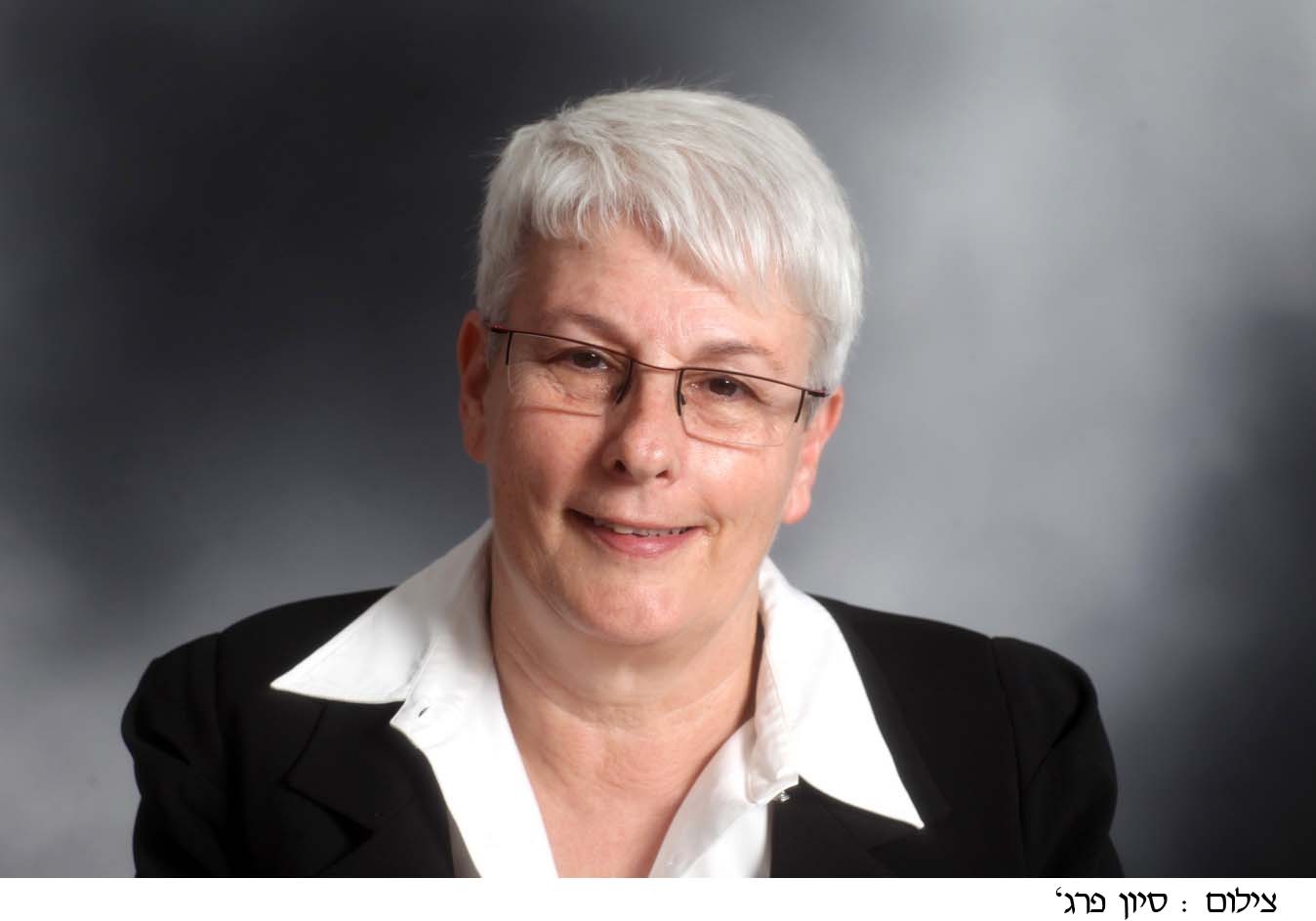 Orna Berry with short white hair and glasses is smiling slightly. She is wearing a black blazer over a white collared shirt. The background is a blurred gray gradient. 