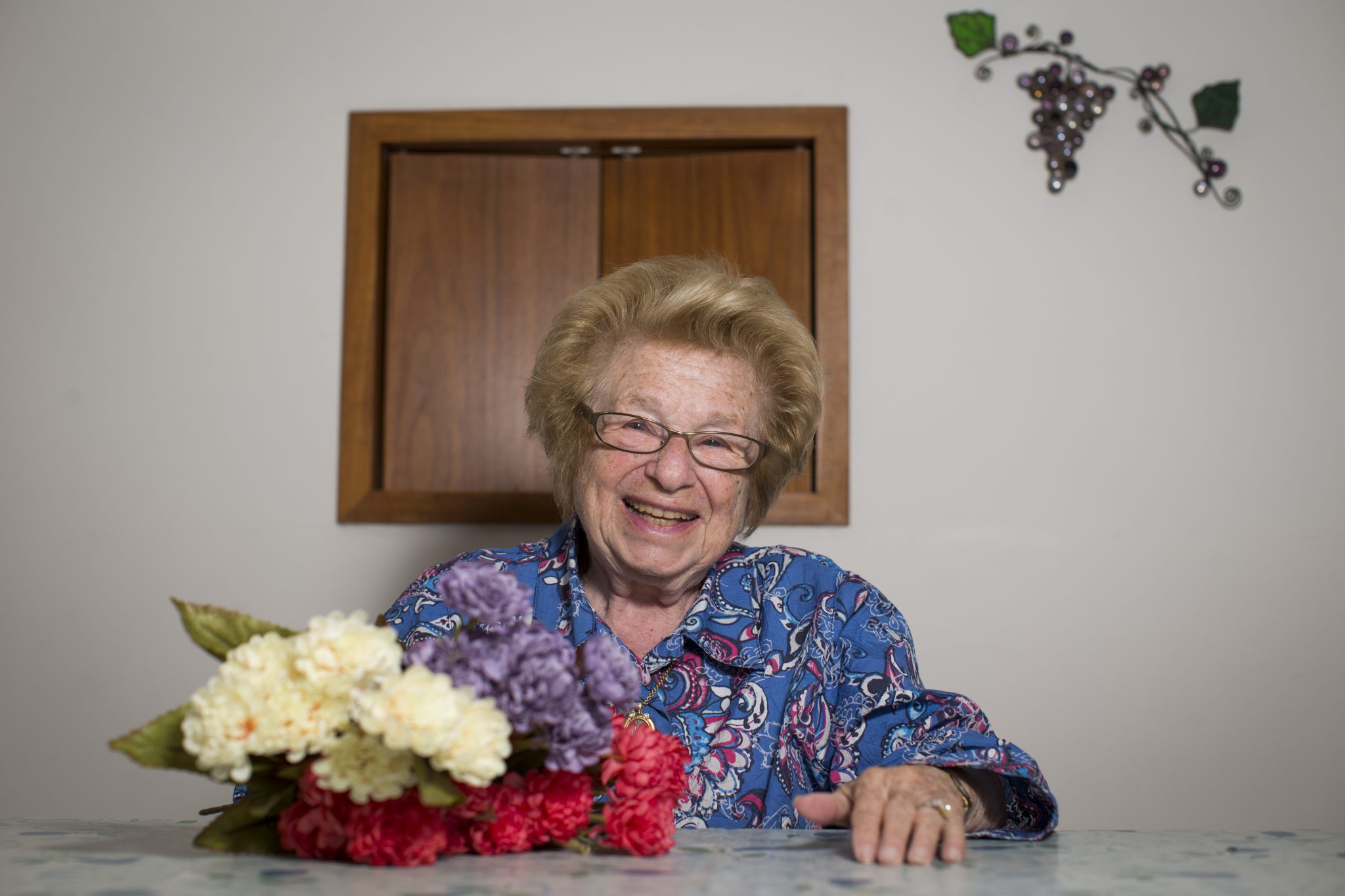 A photo of Dr. Ruth with glasses and a floral blouse, smiling while sitting at a table. In front of her is a colorful bouquet of flowers. A wooden cabinet and metal grapevine decoration are on the wall behind her. 