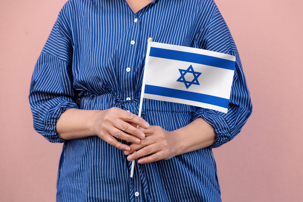 A person dressed in a blue and white striped shirt holds a small Israeli flag with the Start of David against a plain pink background.