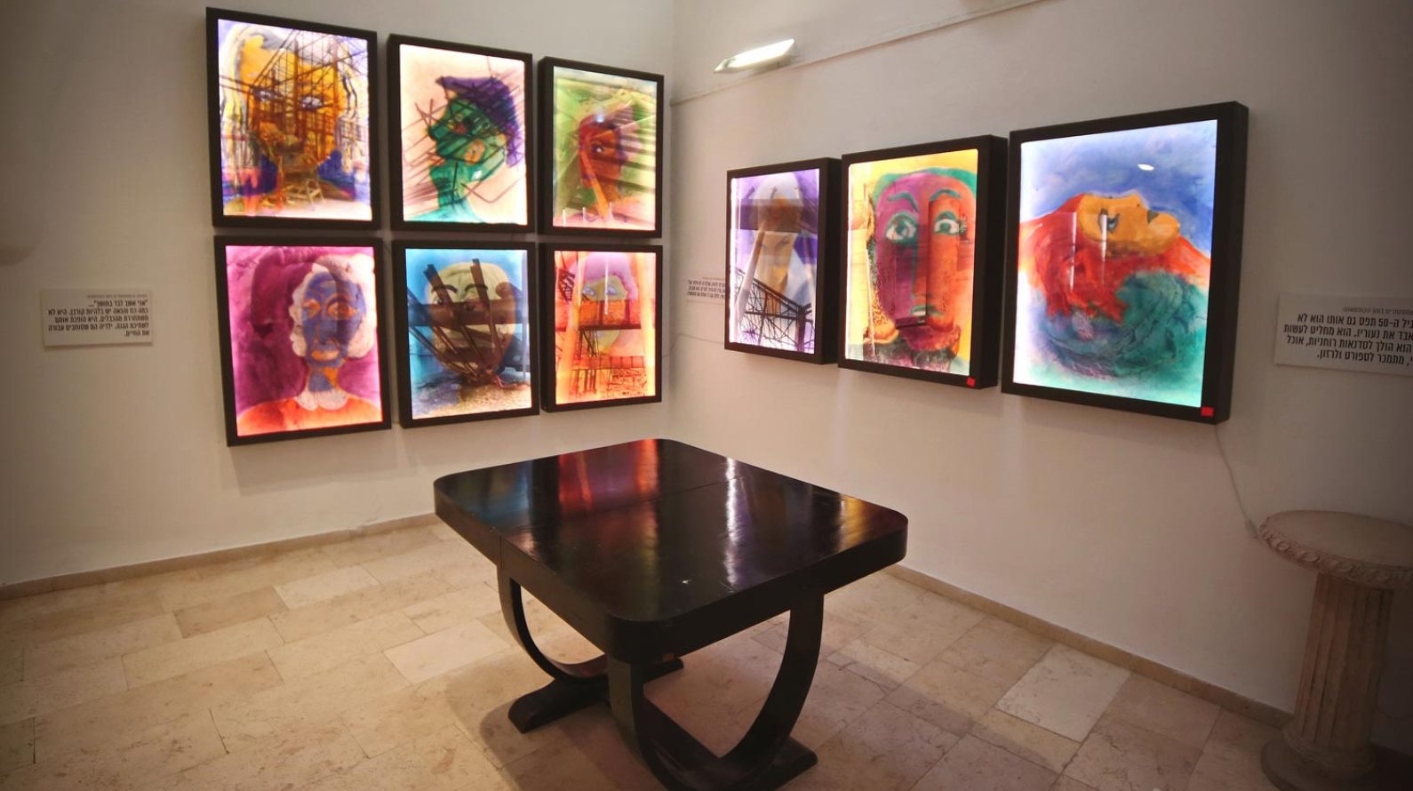 A well-lit gallery displays a dark wooden table in the center, surrounded by vibrant abstract paintings with vivid colors displayed in light boxes on the walls. The artworks have expressive faces and shapes, creating a lively and artistic ambiance.