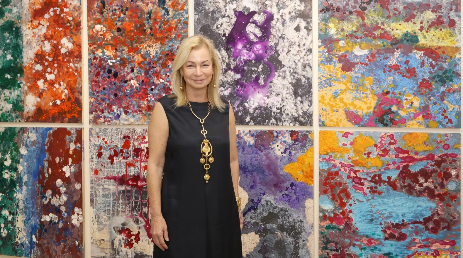 Israeli painter Ariela Wertheimer, with blonde hair in a black dress, stands smiling in front of a vibrant, abstract art display with multiple colorful panels featuring splatters and swirls.
