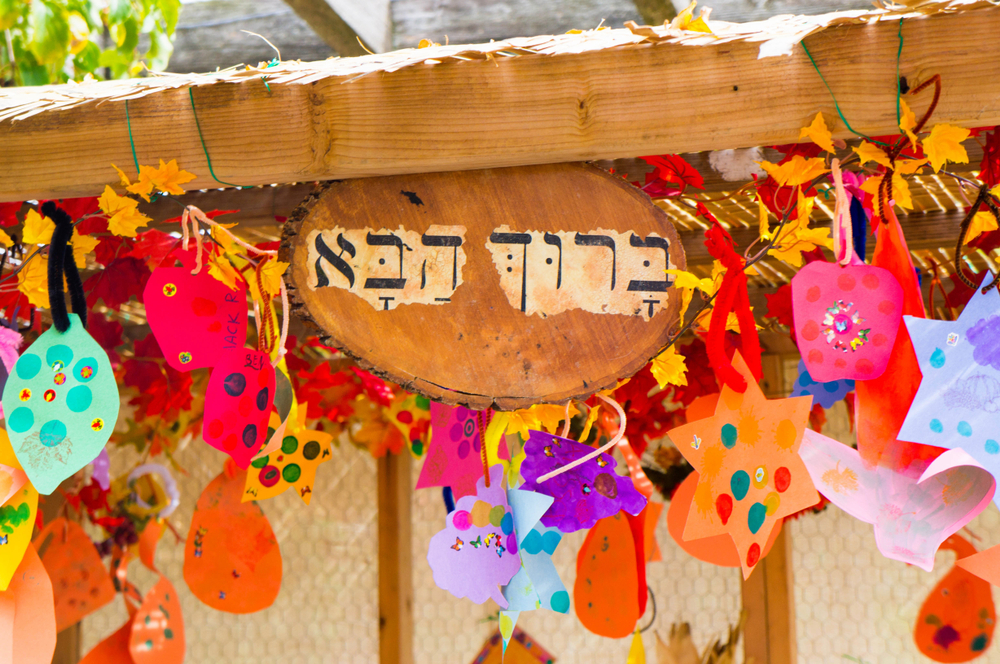 Handmade sukkah decorations are cherished items in Israel. Photo by Liatsh via Shutterstock.com A wooden sign with Hebrew letters hangs on the roof of a Sukkah, surrounded by colorful paper decorations and autumn leaves, creating a festive atmosphere.
