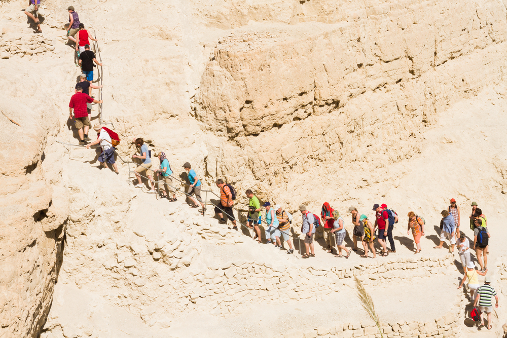 The holiday season is perfect for hiking. Unfortunately the whole of Israel thinks the same. Photo by Shutterstock A group of people, some with backpacks, climb a narrow, winding path along a rocky, sunlit cliff in a desert landscape. They move in single file, holding onto a handrail for support.
