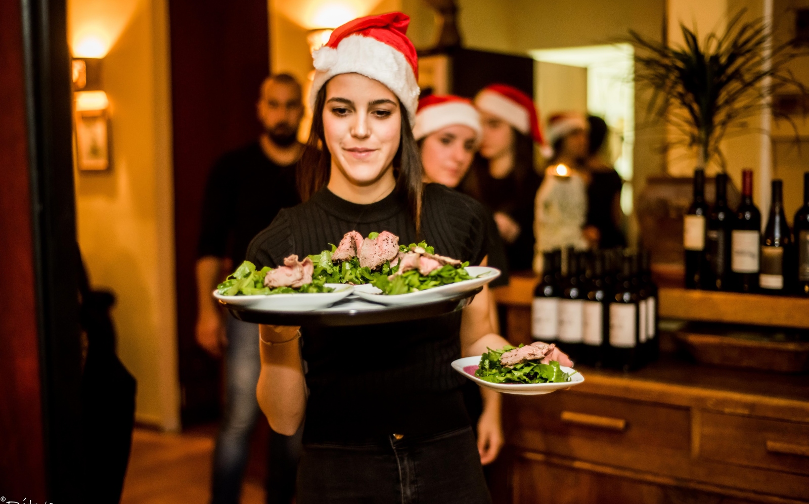A woman wearing a Santa hat serves plates of salad and meat in a festive restaurant setting. Other people in Santa hats and bottles of wine are visible in the background.