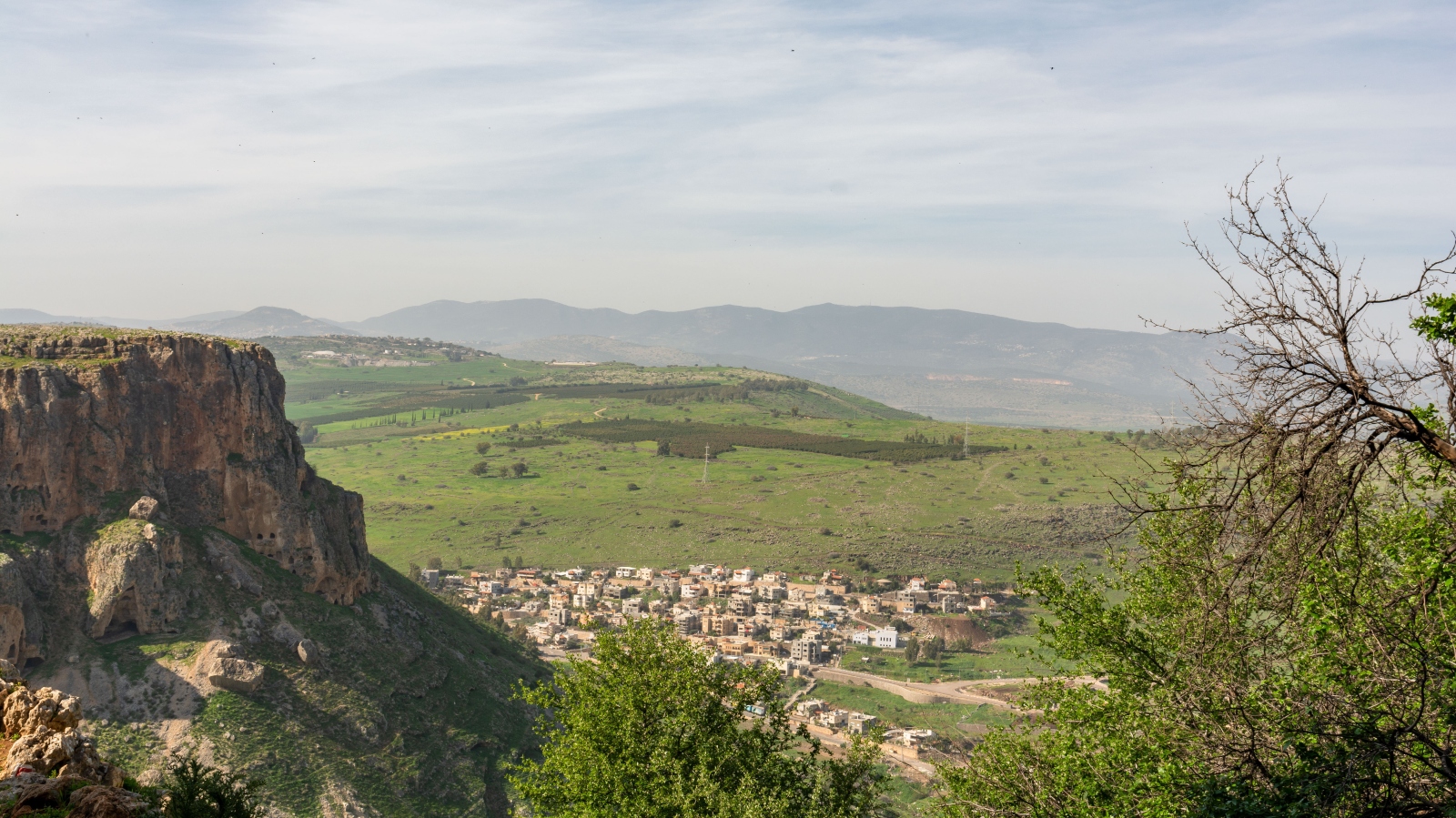 A small village with beige buildings sits at the base of a rocky cliff, surrounded by lush green fields and distant hills under a partly cloudy sky. Sparse trees frame the scene in the foreground.