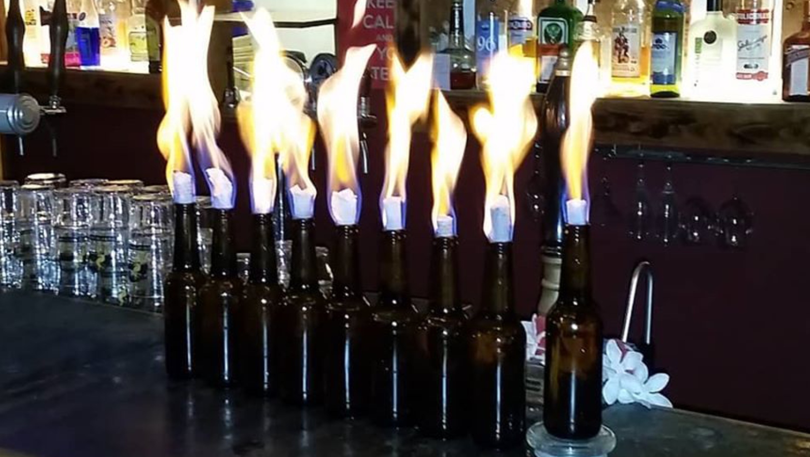 Several glass bottles are lined up on a bar counter as a make-shift, each with a paper-topped neck that is set ablaze, producing bright flames. Shelves of bottles and glasses are visible in the background.