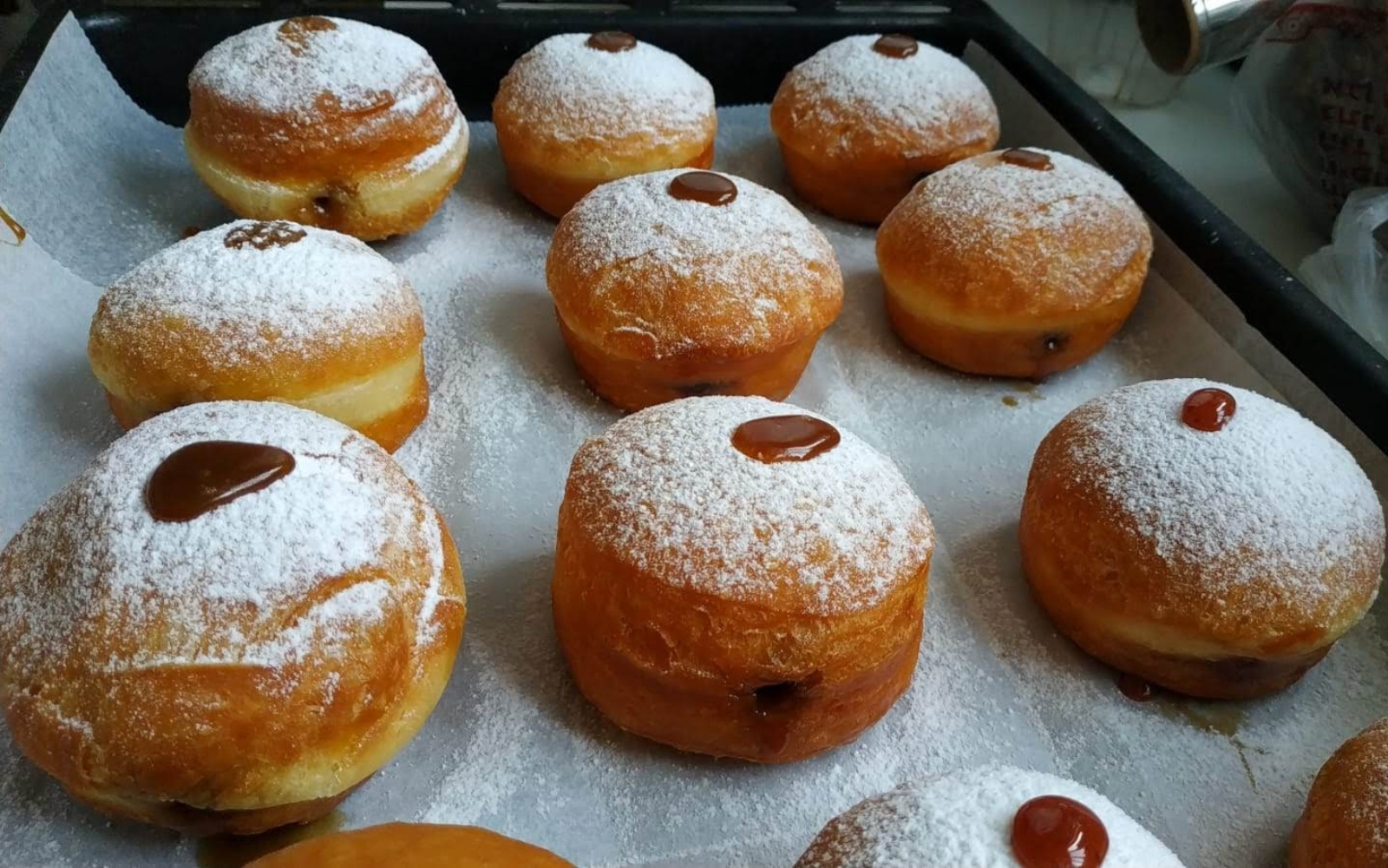 Powdered sugar-covered jelly doughnuts are arranged on a baking tray lined with parchment paper. Some have a spot of jelly or chocolate filling visible on top.