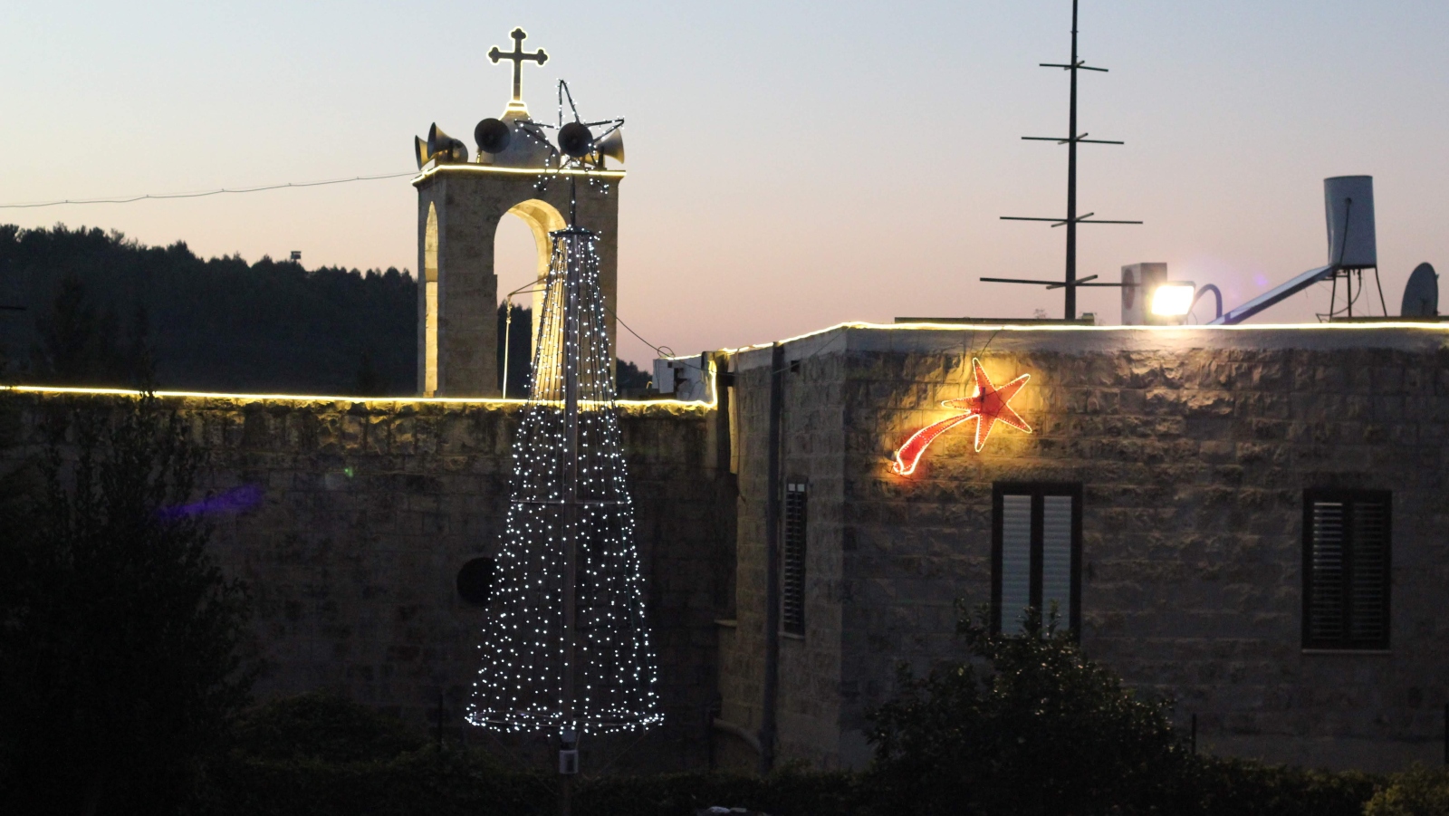 A stone building decorated with holiday lights at dusk, including a lit artificial Christmas tree and a bright shooting star, with trees and a bell tower in the background.