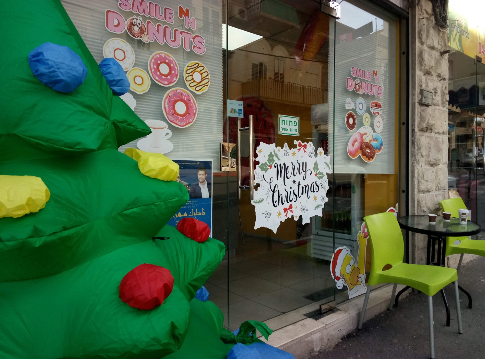 A donut shop with a glass door decorated with colorful donut stickers and a Merry Christmas sign. A large inflatable Christmas tree and green chairs with a small table are outside the entrance.