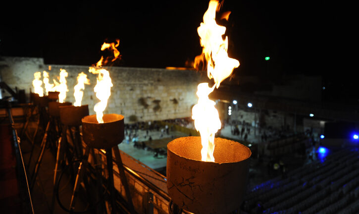 A large menorah burning brightly at night with flames rising, set against the backdrop of the Western Wall in Jerusalem, with people gathered below in the plaza.