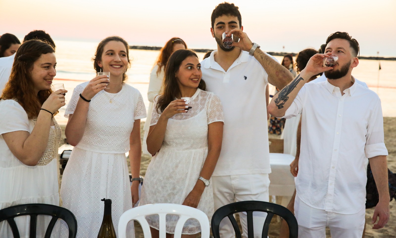 A group of five young adults dressed in white stand on a beach at sunset, smiling and drinking from cups. Plastic chairs and a bottle are visible in the foreground.