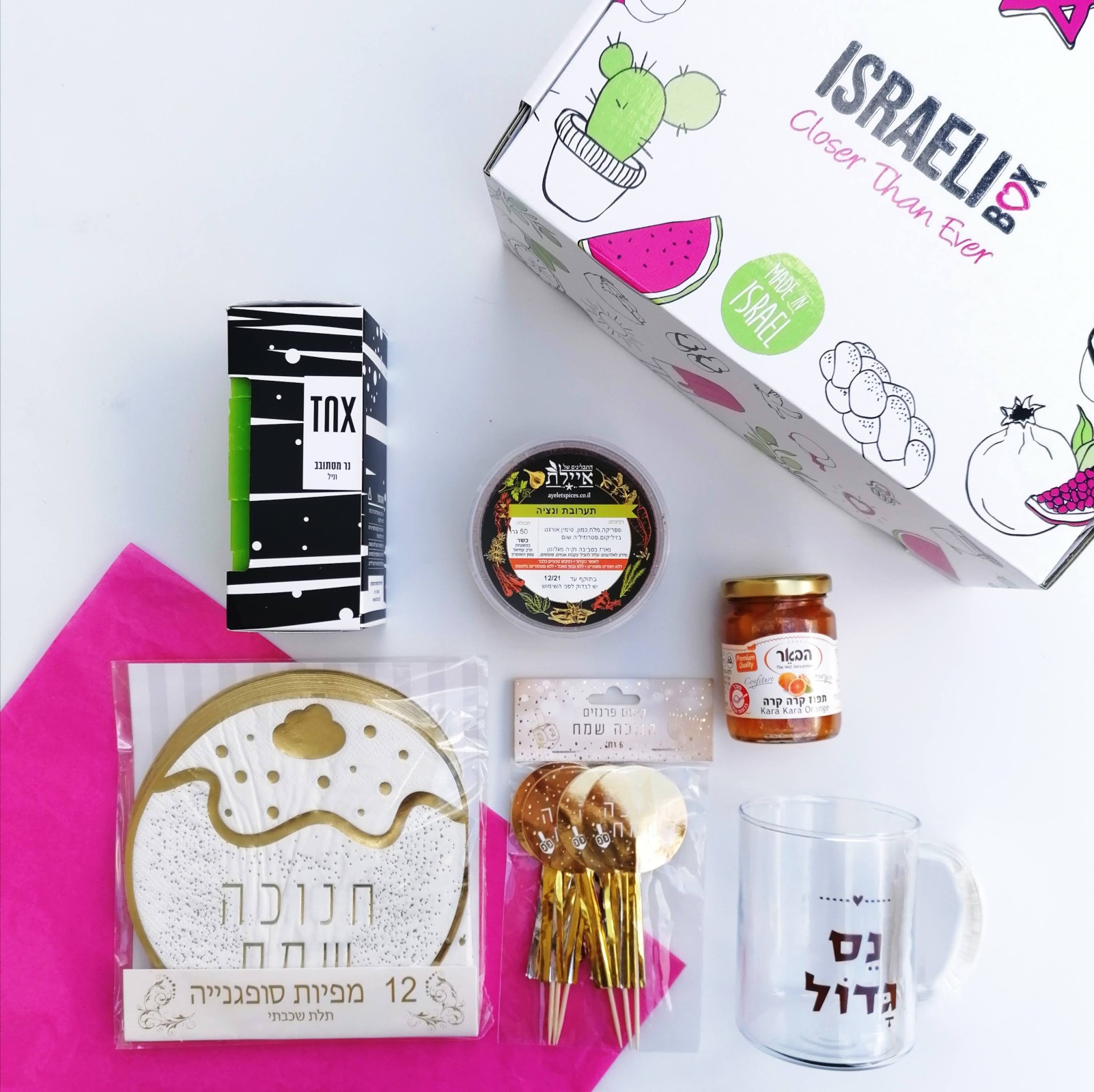 A colorful Israeli gift box with illustrated fruit and cacti, surrounded by festive items: cake topper, striped box, glass mug, jam jar, sweet pastry, and pink tissue paper, all arranged neatly on a white background.