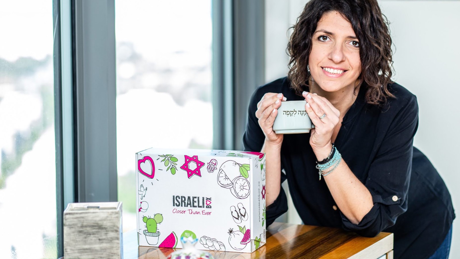 A woman with curly hair smiles while holding a small box with Hebrew writing. Next to her is a colorful box labeled ISRAELI Color Skin Fun decorated with hearts, stars, and cactus illustrations.
