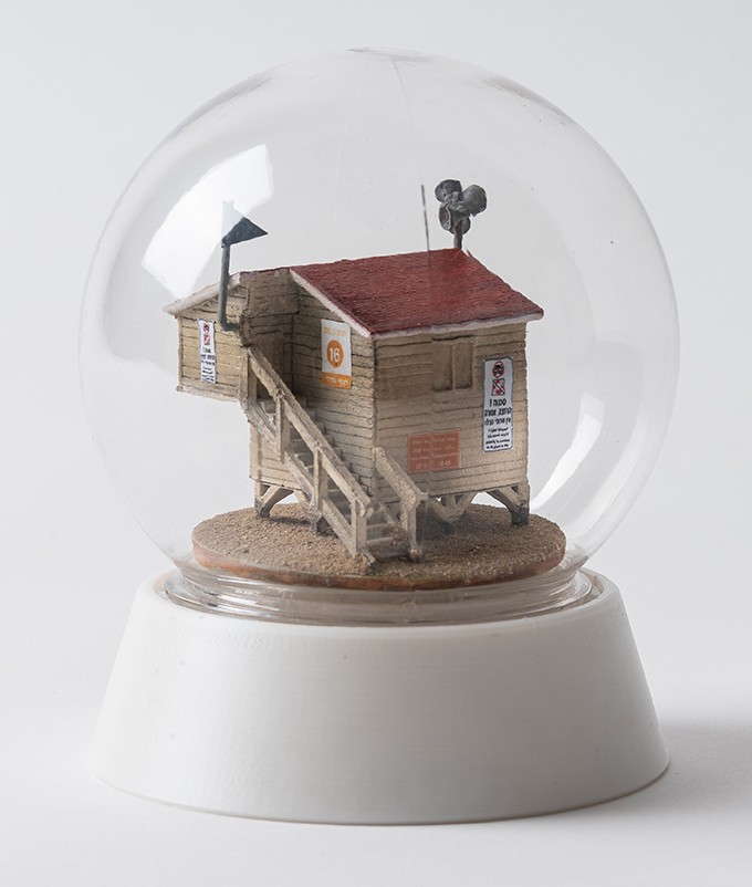 A miniature lifeguard shack with a red roof and signs sits on sand inside a clear snow globe, mounted on a white base.