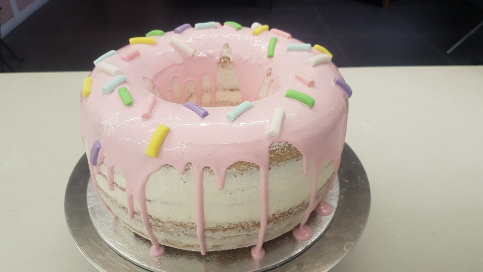 Celebrate the holidays with this beautiful donut cake. Photo still from film A round, frosted donut cake with a pink glaze dripping down the sides, decorated with large, colorful rectangular sprinkles on top, sits on a metallic cake stand.