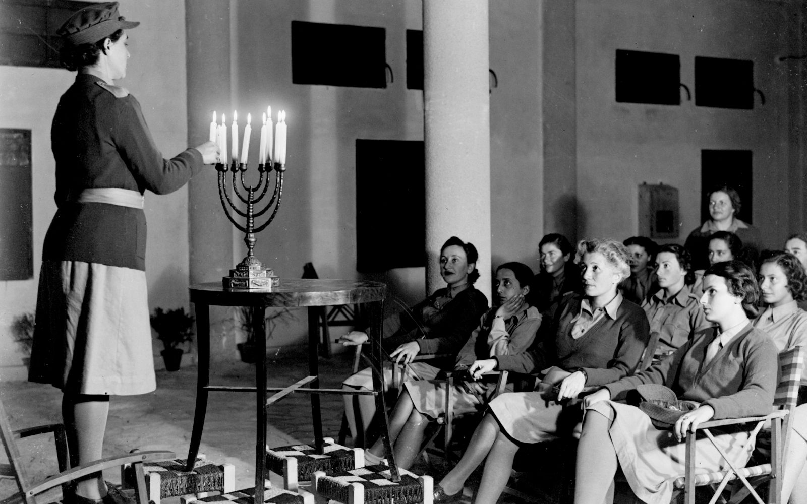 A woman in uniform lights a menorah while a group of women in similar uniforms sit and watch attentively,participating in a Hanukkah celebration in a historic or military setting.