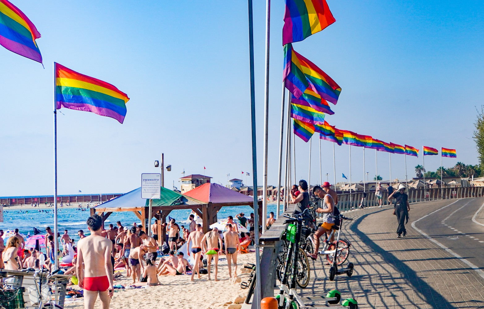 Crowded Hilton beach with people relaxing under colorful umbrellas. Rainbow Pride flags line the boardwalk, and bicycles are parked nearby. The sky is clear, and the ocean waves gently reach the shore.