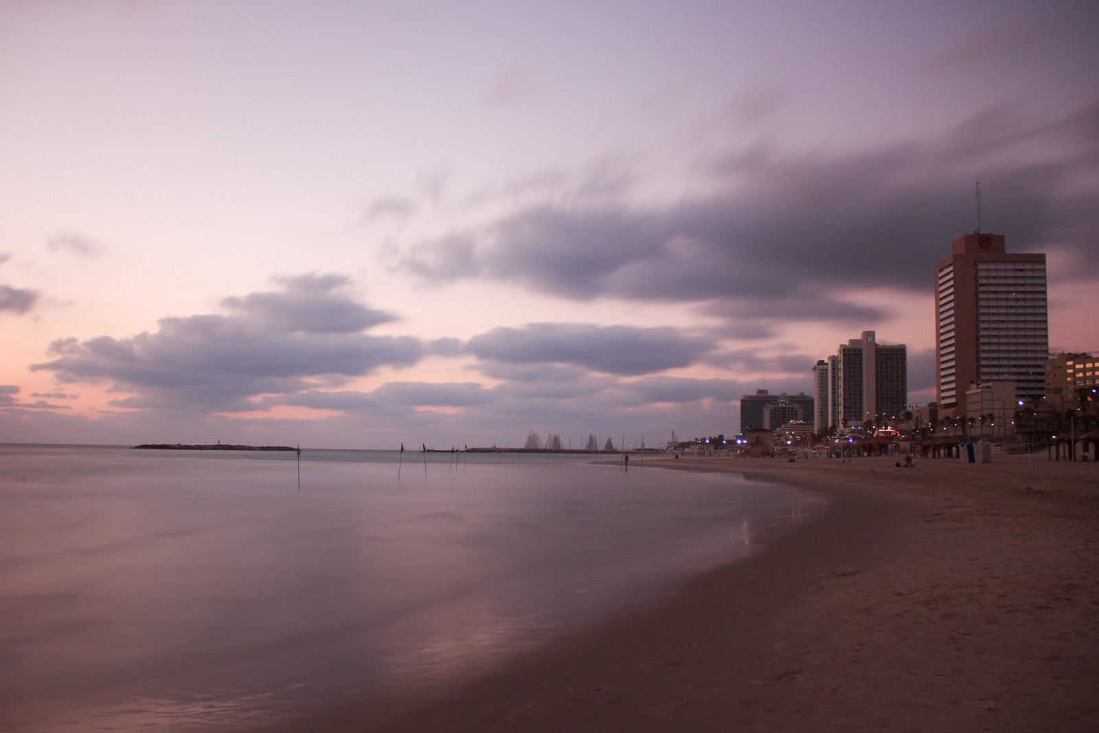A tranquil view of Banana Beach at dusk with a pastel-colored sky and scattered clouds. The calm sea reflects the subtle hues. Tall buildings line the right side, with some lights starting to illuminate. Minimal people are present on the sandy shore.