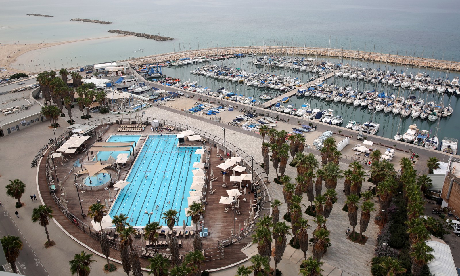 Aerial view of the coastal marina near Gordon Beach with numerous docked boats. In the foreground, theres the large outdoor Gordon swimming pool surrounded by palm trees and deck chairs. The sky is overcast, and a sandy beach is visible in the background.