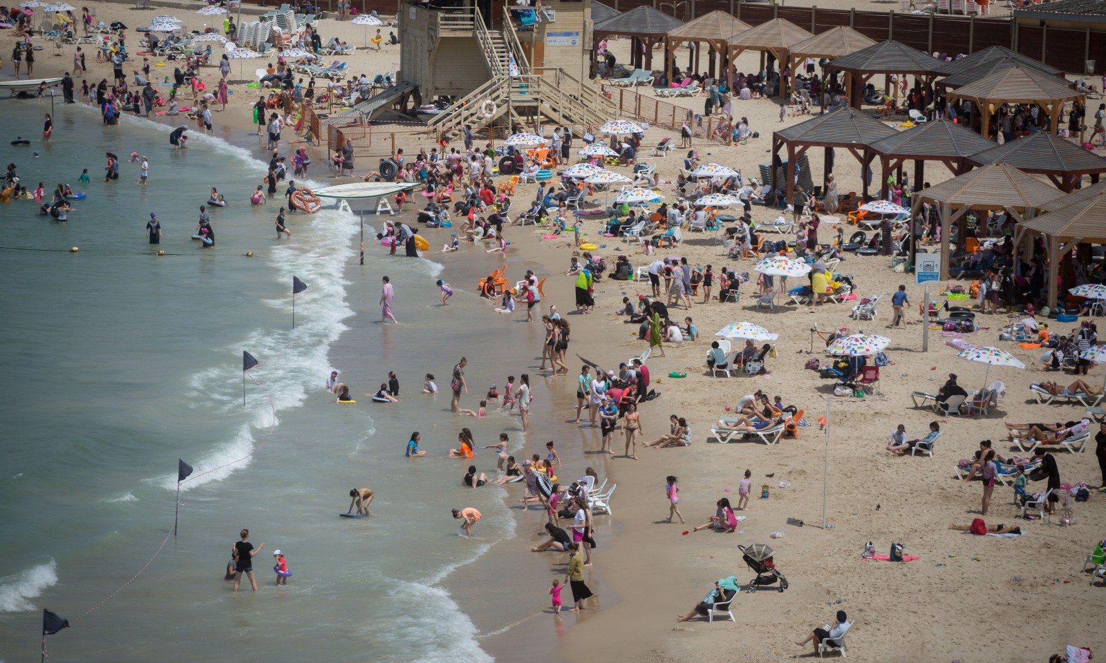 A crowded beach scene with people swimming, playing in the sand, and relaxing under sunshades. Lifeguard tower and wooden pergolas are visible in the background. The shoreline is busy with beachgoers enjoying a sunny day.