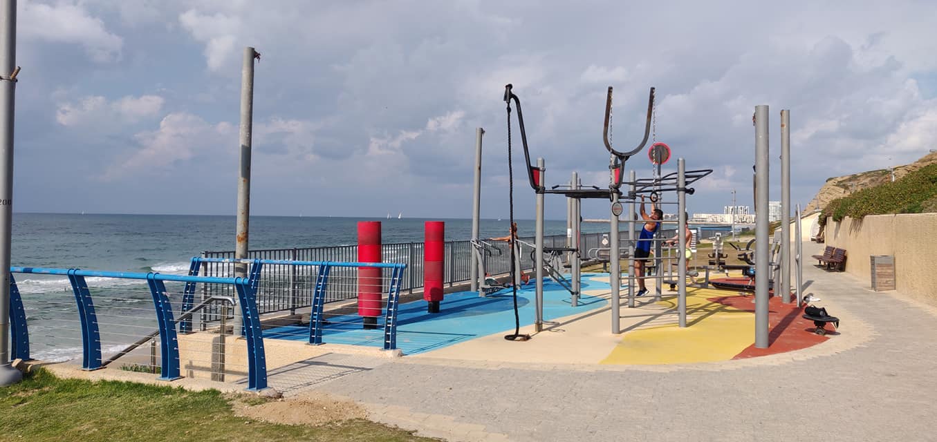 Outdoor fitness equipment near a beach. Various exercise structures are set on a colorful rubber surface. The ocean and clouds are visible in the background, along with a pathway. A person is using the equipment.
