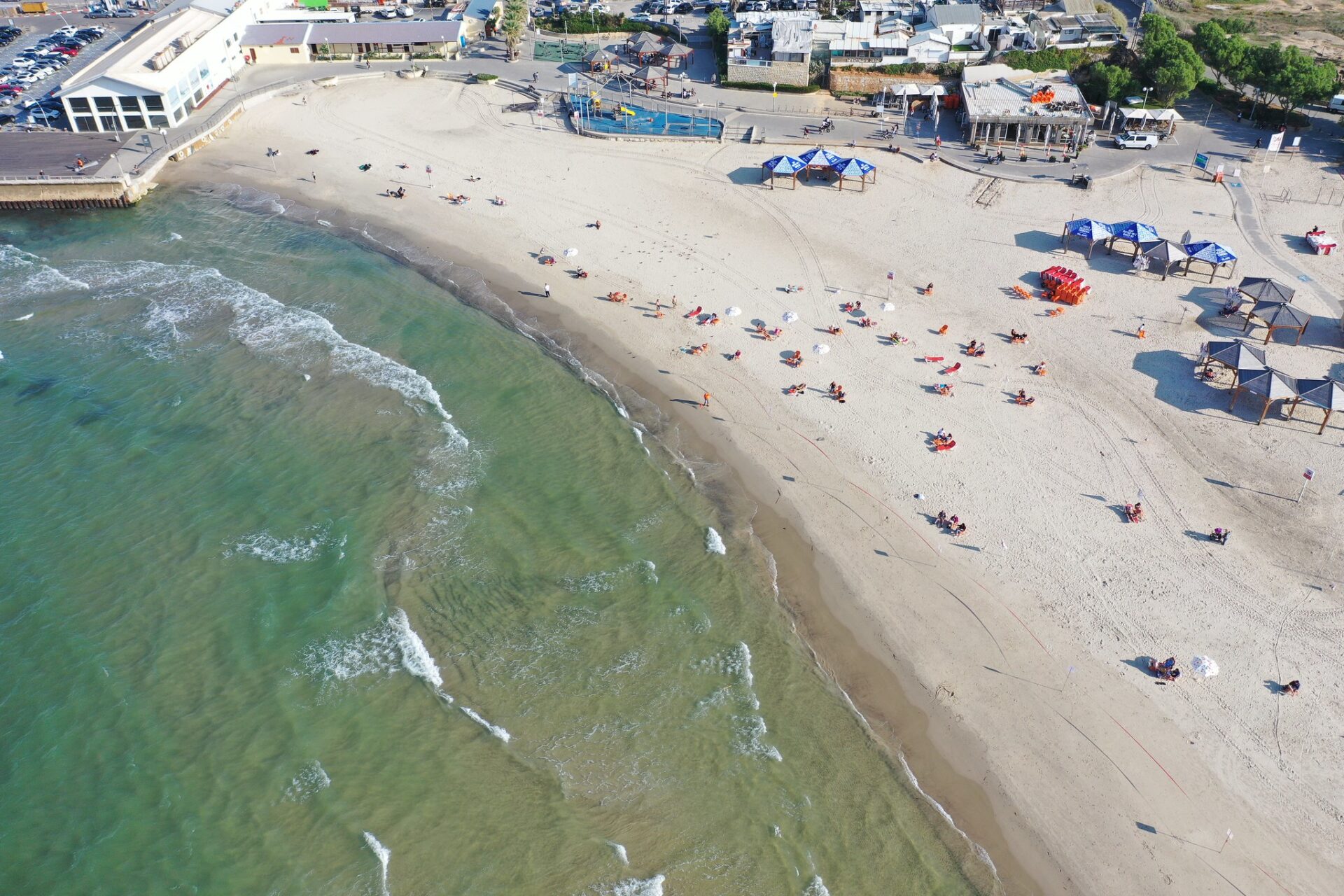 Aerial view of Metzitzim Beach, with gentle waves and a sandy shore. People are lounging under blue umbrellas and red chairs scattered across the sand. Buildings are visible at the top, and the sea is on the left.
