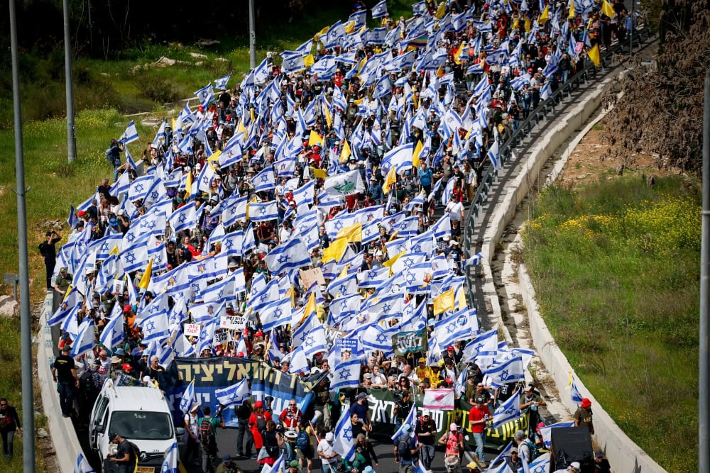 A large crowd of people march along a road, carrying numerous Israeli flags and banners, demonstrating a peaceful protest on a sunny day. 